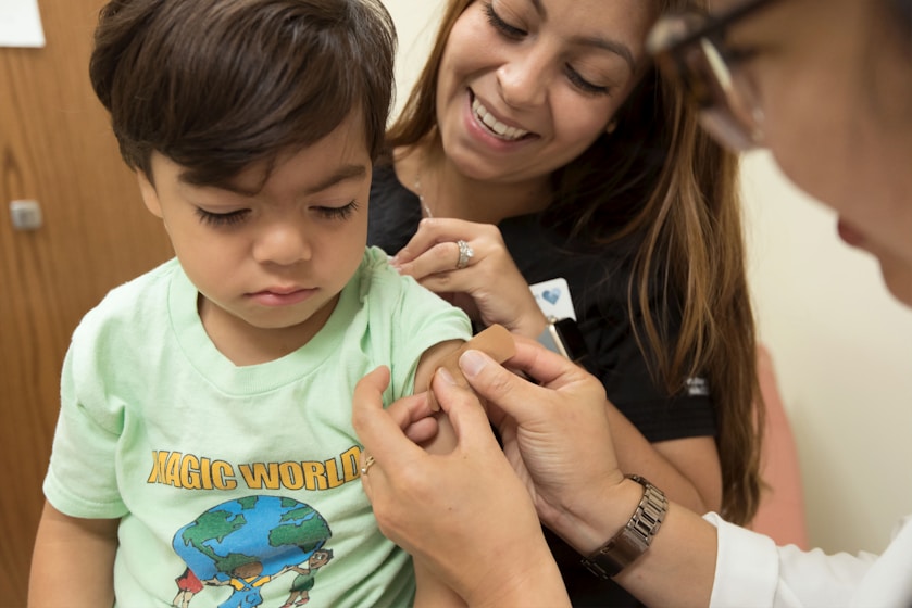 A boy receiving a bandage from a smiling nurse while his mother watches