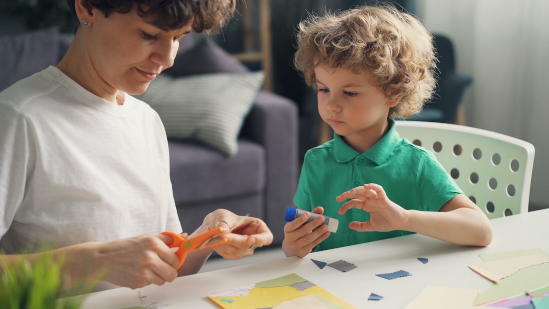 A mother helping her son with a craft activity using scissors and glue