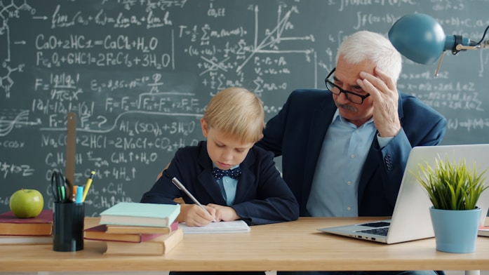 A boy writing at a chalkboard desk with an elderly tutor