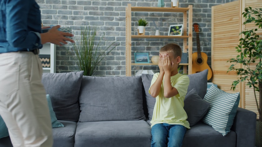A boy covering his face while sitting on a grey couch