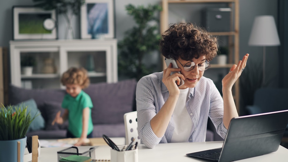 A woman on the phone at her laptop while a child plays nearby