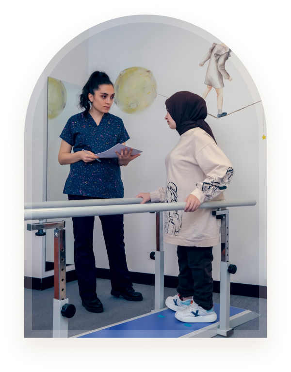 A therapist holding a clipboard while assisting a young woman using parallel bars in a rehabilitation room at Hiba by Anthalora